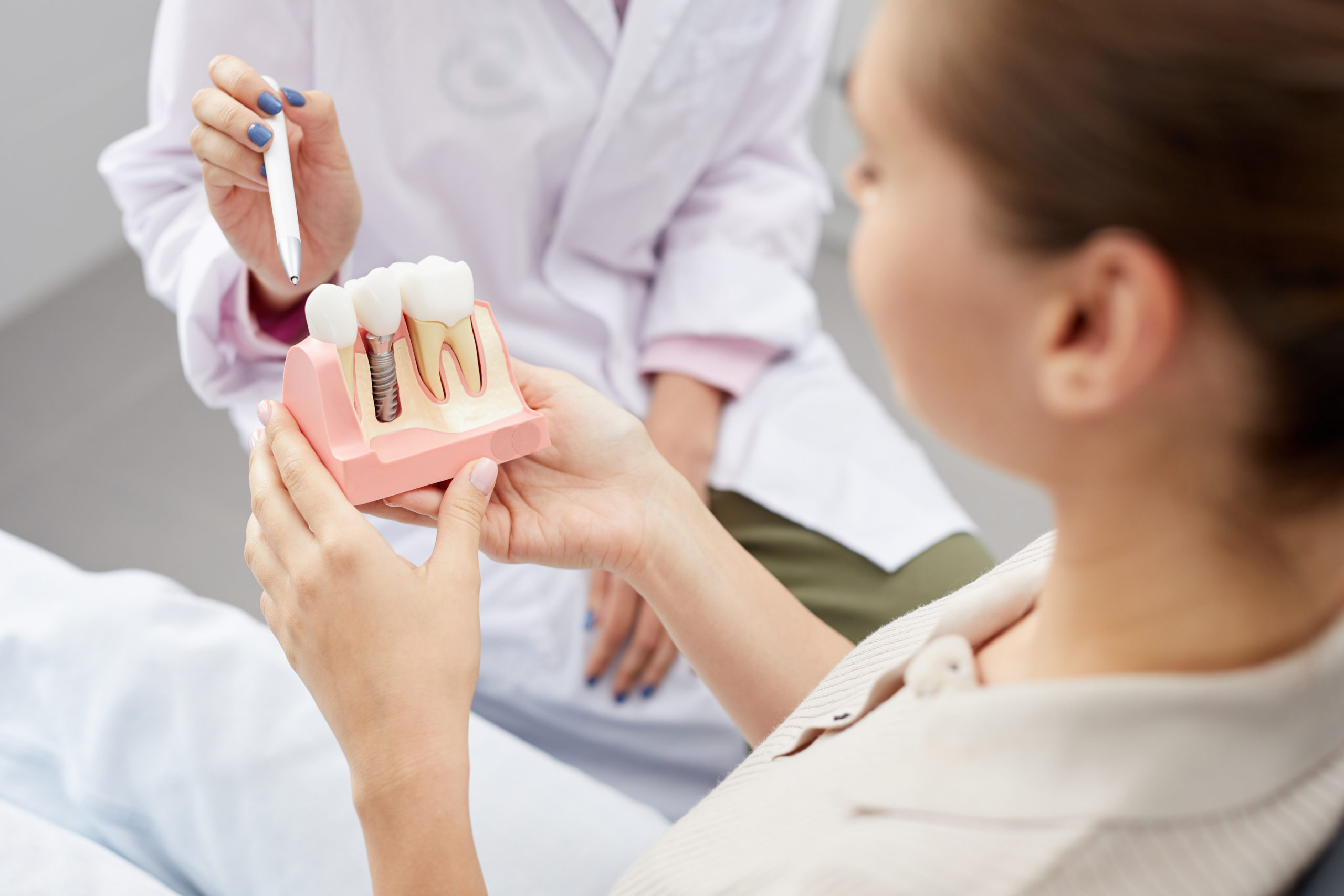 Eine weibliche Zahnärztin zeigt auf ein Zahnimplantat-Modell, welches eine Patientin in den Händen hält und sich anschaut.